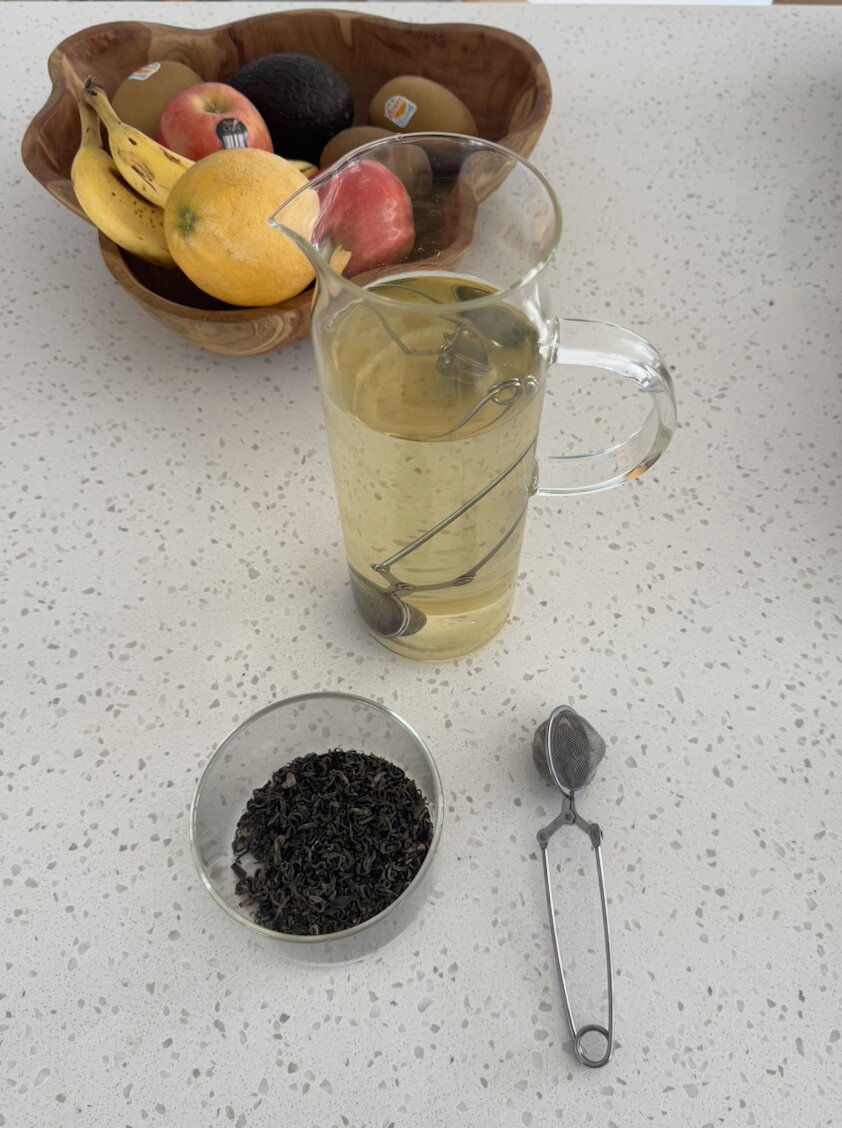 Loose-leaf green tea in a glass bowl, a stainless steel snap-ball infuser, and a two-litre glass jug of pale gold cold brew on the kitchen bench